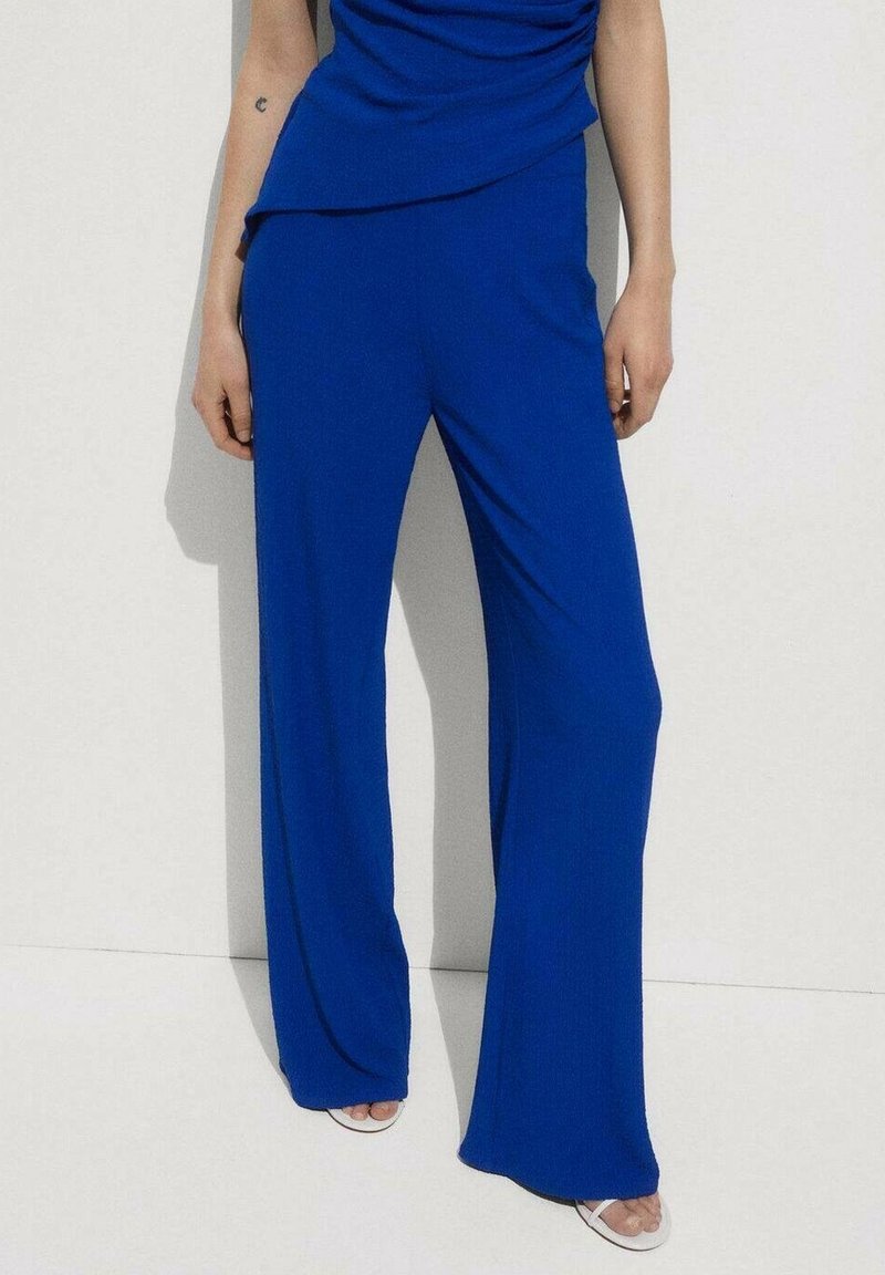 Femme portant un pantalon large bleu vif et un haut assorti, se tenant devant un mur blanc, avec des sandales ouvertes blanches visibles.