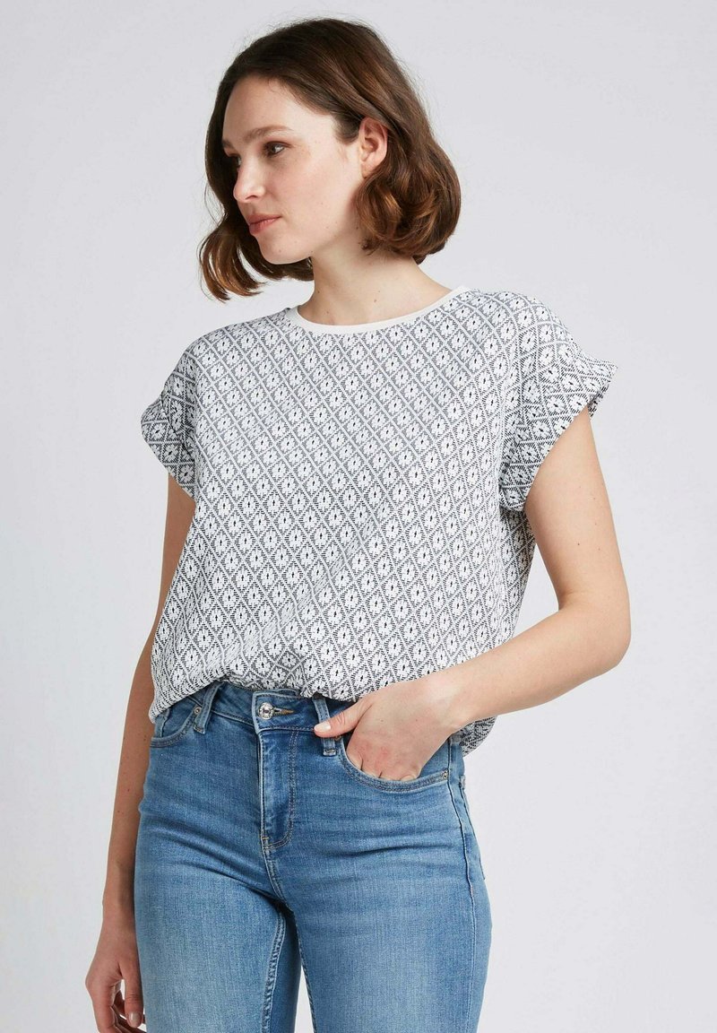 Blouse blanche à manches courtes avec un motif en diamond noir, décolleté rond et coupe ample, associée à un jean bleu pour un look décontracté.