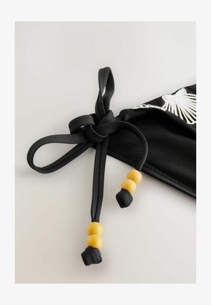 Czarna tkanina z węzłem związanym z żółtymi drewnianymi koralikami. Węzeł zabezpiecza odzież, ozdobioną białą haftowaną krawędzią.