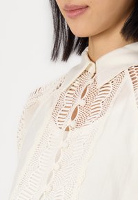 Witte blouse met intrigerende kantdetails, een knoopsluiting en een kraag. De stof lijkt glad met een zachte textuur.