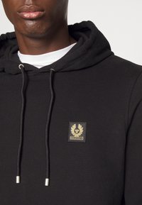 Musta huppari, joka on valmistettu puuvillaseoksesta. Siinä on kiristysnauhalla varustettu huppu, resorit hihansuissa ja kultainen logopläjäys rinnassa brändäystä varten.