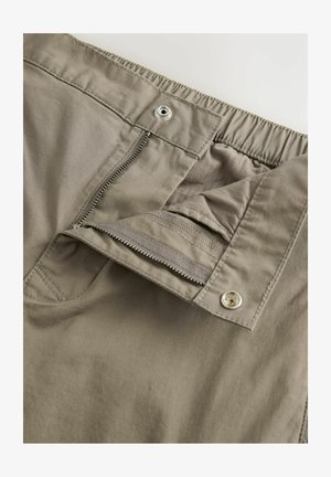 Lys grå bukser med lynlåse foran, elastisk talje og en glat tekstur, med en metal trykknaplukning i taljen.