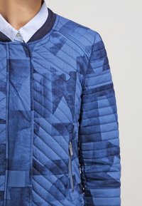Blaue gesteppte Jacke mit geometrischen Mustern, ausgestattet mit einer Reißverschlusstasche und einem gerippten Kragen. Das Material hat eine glatte Textur und Farbverläufe.