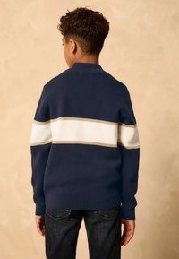 Tummansininen neulepaita, jossa vaaleanbeiget ja valkoiset vaakaraidat. Varustettu resorikauluksella ja -hihoilla. Yhdistetty tummiin farkkuihin.