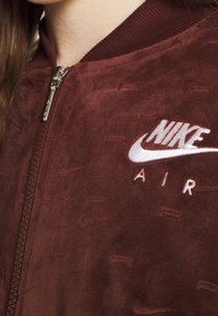 Burgundská semišová bomber bunda Nike Air, se zipovým zapínáním a bíle vyšitým logem. Hladký límec a jemný texturový detail.