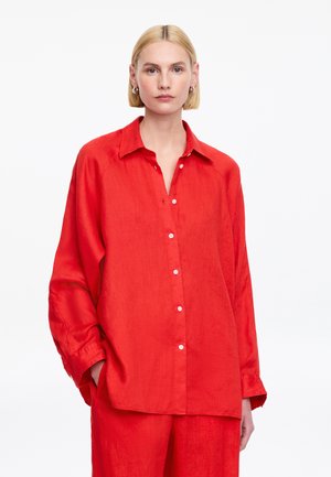 Mujer rubia con cabello corto que lleva una camisa roja de botones holgada y pantalones rojos a juego, de pie contra un fondo blanco.