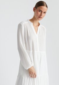 Robe blanche à manches longues en tissu léger texturé. Elle présente un col henley, un design à volants et de légères manches bouffantes.
