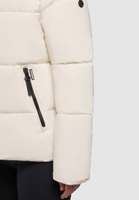 Steppjacke in Off-White, mit gesteppten Abschnitten, glatter Textur und einer Reißverschlusstasche am linken Ärmel mit schwarzem Metall.