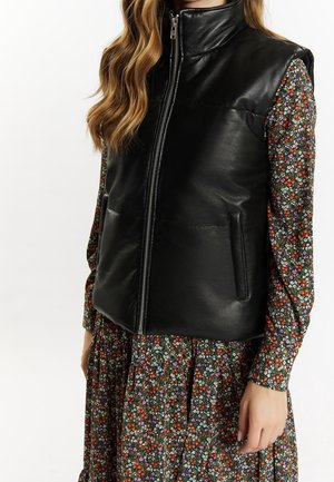 Femme portant un gilet en cuir noir zippé sur une robe à manches longues à fleurs avec de petites fleurs rouges, blanches et violettes.