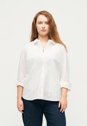 Woman with long red hair wearing a white button-up blouse with floral embroidery and dark blue jeans, standing against a plain background.