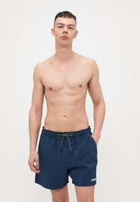 Marine svømmebukser laget av lett stoff, med elastisk midje og snor, samt en liten hvit logo på venstre side.