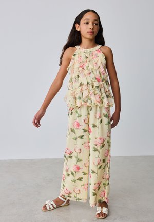 Djevojka u kremastom cvjetnom topu bez rukava sa volanima i odgovarajućim širokim hlačama, stoji na sivom podu nasuprot sivom zidu.