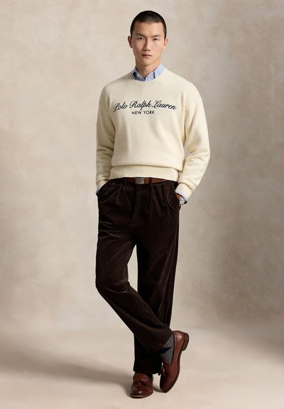 Άνδρας που φοράει κρεμ πουλόβερ Polo Ralph Lauren, ανοιχτό μπλε πουκάμισο με γιακά, σκούρο καφέ παντελόνι κορδόνι και καφέ δερμάτινα παπούτσια, στέκεται με τα χέρια στις τσέπες.