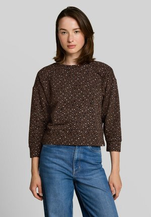 Kobieta o brązowych włosach do ramion, ubrana w brązową bluzkę w long sleeve w panterkę i niebieskie dżinsy, stojąca na jednolitym tle.