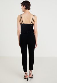 Černý smocked top s tenkými ramínky a volánkovým lemem, kombinovaný s přiléhavými černými kalhotami a páskovými černými vysokými podpatky s detaily.