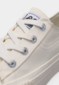 Κοντινό πλάνο ενός λευκού πάνινα παπουτσιού με λευκά κορδόνια, μεταλλικά οφθαλμούς με τη μάρκα "Lee 1889" και εμφανές υφασμάτινο λογότυπο στη γλώσσα.