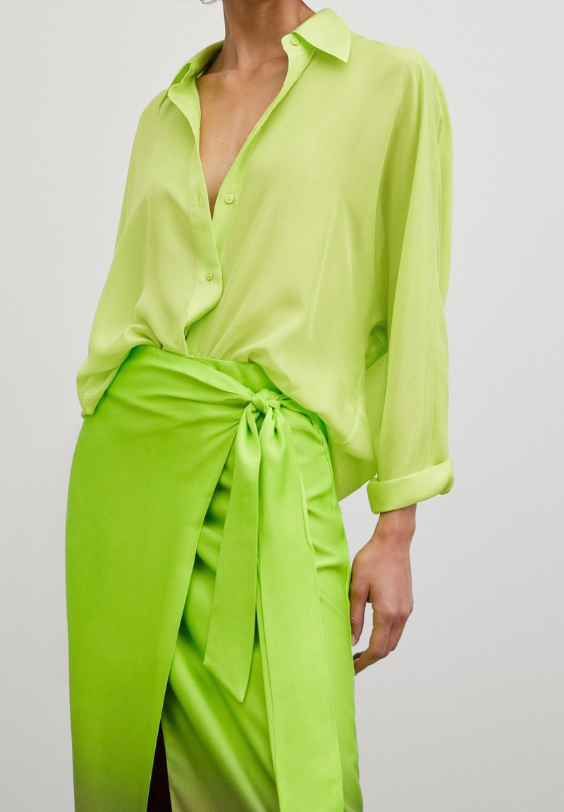 Femme portant un chemisier à manches longues vert citron vif, rentré dans une jupe portefeuille taille haute assortie nouée sur le côté.