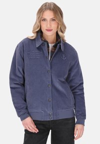 Młoda kobieta z falującymi blond włosami, ubrana w niebieską kurtkę z kordurowej tkaniny na koszulę w kratę i czarne spodnie, pozująca z jedną ręką przy biodrze.