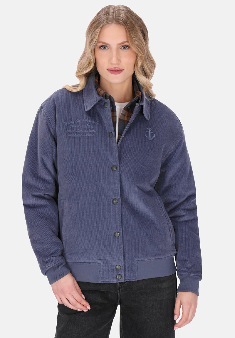 Młoda kobieta z falującymi blond włosami, ubrana w niebieską kurtkę z kordurowej tkaniny na koszulę w kratę i czarne spodnie, pozująca z jedną ręką przy biodrze.