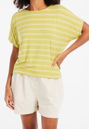 Femme portant une chemise à manches courtes rayée vert clair et un short beige, debout avec une main sur la hanche.