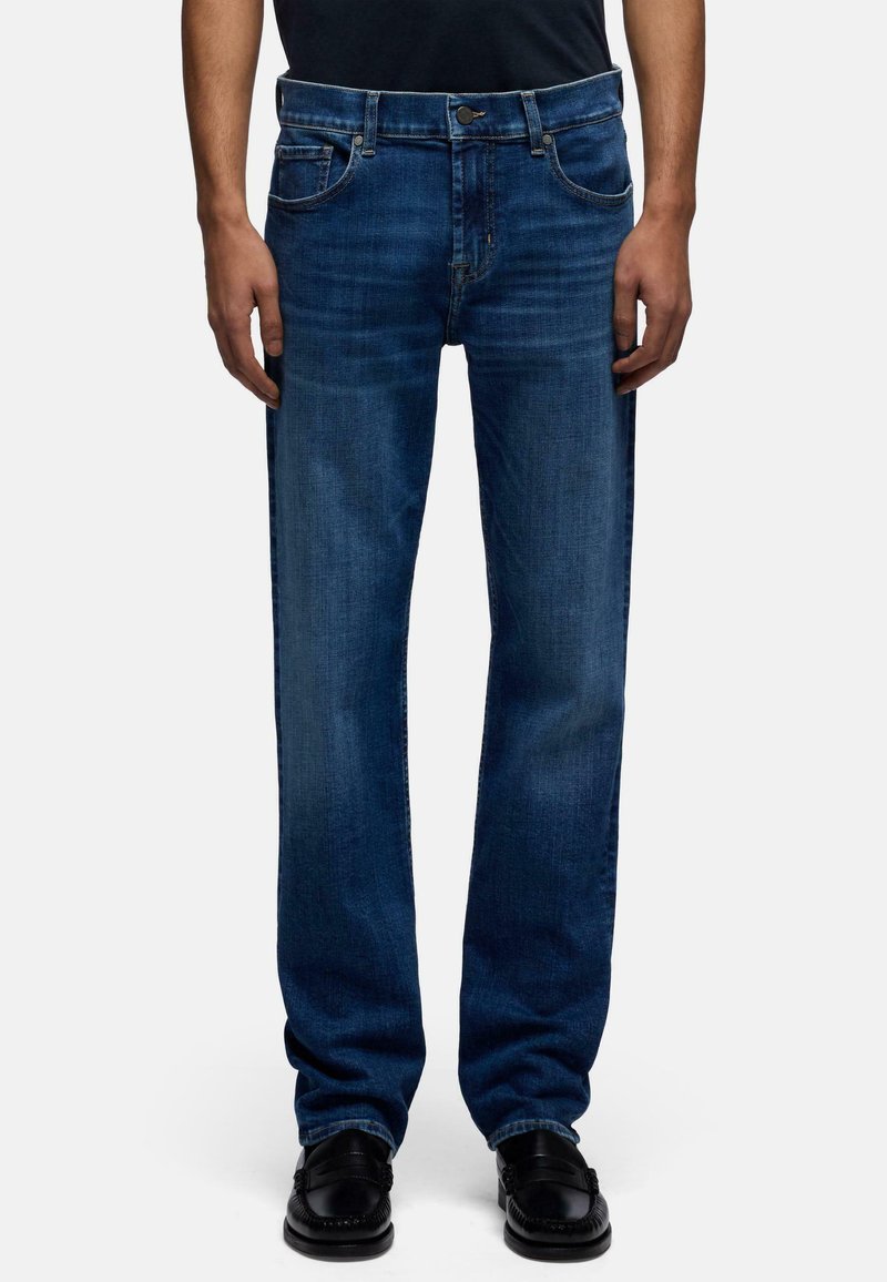Uomo che indossa jeans blu a gamba dritta e mocassini in pelle nera, in piedi su uno sfondo chiaro e uniforme.