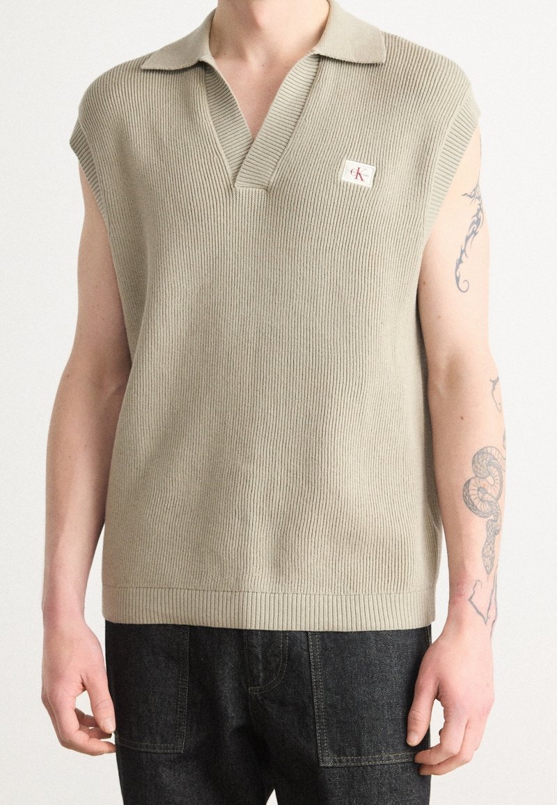 Veste sans manches en tricot beige avec col et petite étiquette logo portée par une personne avec des tatouages sur le bras droit, associée à un pantalon foncé.