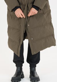 Olivgrüner Puffermantel mit Reißverschluss und Druckknöpfen, ausgestattet mit einem gesteppten Design. Trägt man über schwarzen schmalen Hosen und schwarzen Knöchelstiefeln.