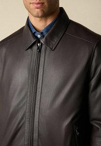 Muž v hnědé kožené zipové bundě přes modrou košili s límečkem, který vykukuje z límečku a horní části hrudníku.