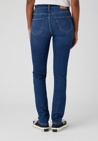 Jeans in denim skinny di colore blu scuro, caratterizzati da un alto punto vita, due tasche posteriori con dettagli di cucitura e una texture sottile. Abbinati a sneakers nere.