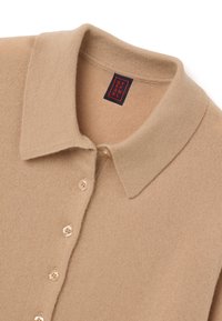 Beige gebreide trui met een klassieke kraag, voorzien van vijf knoopsluitingen, textiel met structuur en een label met rode en zwarte letters.