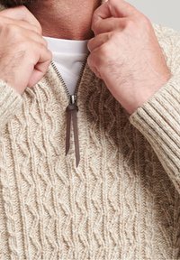 Beige Kabelstrickpullover mit hohem Kragen und Reißverschluss, ausgestattet mit einem Lederzieher. Der Stoff wirkt strukturiert und hat eine weiche Oberfläche.