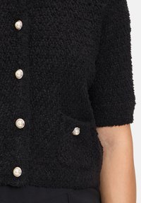 Schwarze strukturierte Strickjacke mit kurzen Ärmeln, ausgestattet mit perlenähnlichen Knöpfen und einer seitlichen Tasche mit dekorativem Knopfdetail.