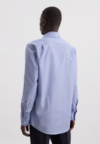 Uomo che indossa una camicia a maniche lunghe a righe verticali blu e bianche, visto di spalle contro uno sfondo chiaro e uniforme.