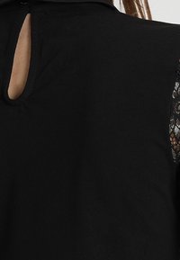 Svart topp med nyckelhålsdetalj i ryggen, korta spetsärmar, mjuk bomullstextur och en avslappnad passform.