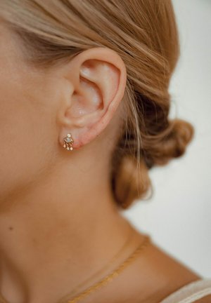 Gold earrings with small white stones, featuring a circular design and three dangling accents, shown on a woman’s ear.