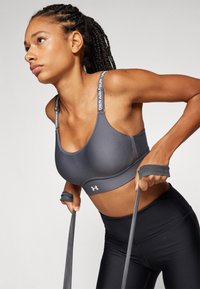 Sujetador deportivo gris con diseño de espalda nadadora, tirantes elásticos y logo de Under Armour; combinado con leggings negros; modelo utilizando bandas de resistencia.