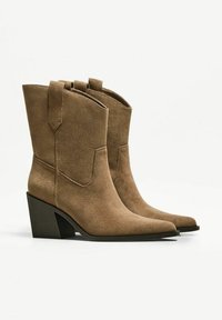 Stivali da cowboy in suede marrone con punta affusolata, tacco impilato di altezza media e linguette sui lati. Tessuto liscio con dettagli minimi.