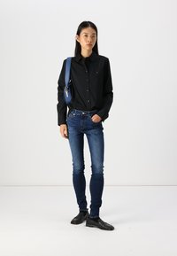 Zwarte overhemdd met lange mouwen, blauwe heuptas, donkerblauwe skinny jeans en zwarte schoenen. Eenvoudig design, gladde textuur.