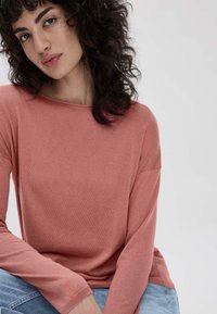 Rosa langärmliges Oberteil mit weitem Ausschnitt und gerippter Textur. Das Modell hat lockige Haare und sitzt lässig. Sie trägt hellblaue Jeans.