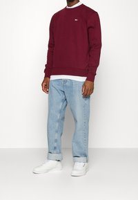 Felpa bordeaux realizzata in morbido materiale con polsini e orlo a costine, indossata sopra una maglietta bianca, abbinata a jeans azzurri dal taglio rilassato e sneakers bianche.