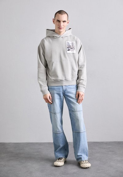 Szary hoodie z kapturkiem i przednią kieszenią, ozdobiony nadrukiem ze zdjęciem, zestawiony z jasnoniebieskimi szerokimi spodniami dżinsowymi oraz obuwiem o fakturze i wzorze.