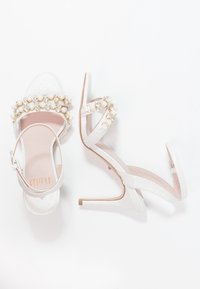 Sandales à talons hauts blanches avec des accents de perles et de bijoux. Design ouvert avec des lanières croisées et une lanière de cheville réglable.