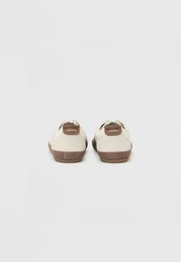 Zapatillas de ante beige con suelas de goma marrón, que presentan un talón trasero texturizado con un parche de logo y un diseño minimalista.