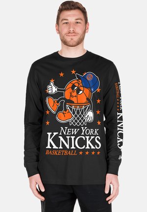 Mann trägt ein schwarzes langärmeliges New York Knicks Shirt mit einem Cartoon-Basketballcharakter und dem Teamnamen auf der Vorderseite und am Ärmel.