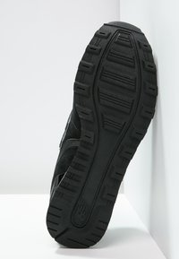 Suola nera di sneaker con un motivo di battistrada profondo inclinata contro una parete e un pavimento bianchi.