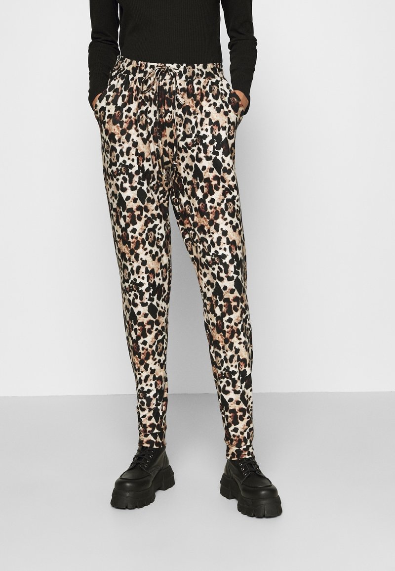 Personne portant un pantalon à cordon en imprimé léopard, un haut noir à manches longues et des bottes à plateforme noires imposantes, se tenant devant un fond simple.