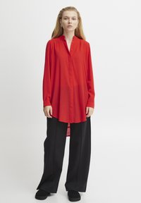 Camicia rossa a maniche lunghe con colletto alla cinese e bottoni. Tessuto fluido, leggermente più lungo sul retro. Indossata con pantaloni neri larghi.