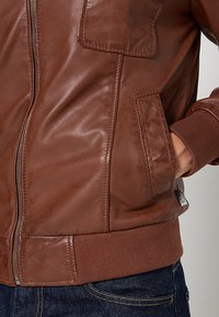 Bruine leren jas met een ritsluiting, twee borstzakken, ribgebreide manchetten en een gladde structuur. Gepersonaliseerde pasvorm met een afgeronde onderkant.