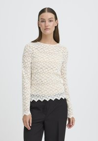 Blonde blonde bluse med lange ærmer i cremefarvet med blomstermønster, bølget kant og en semi-gennemsigtig tekstur, parret med sorte bukser.