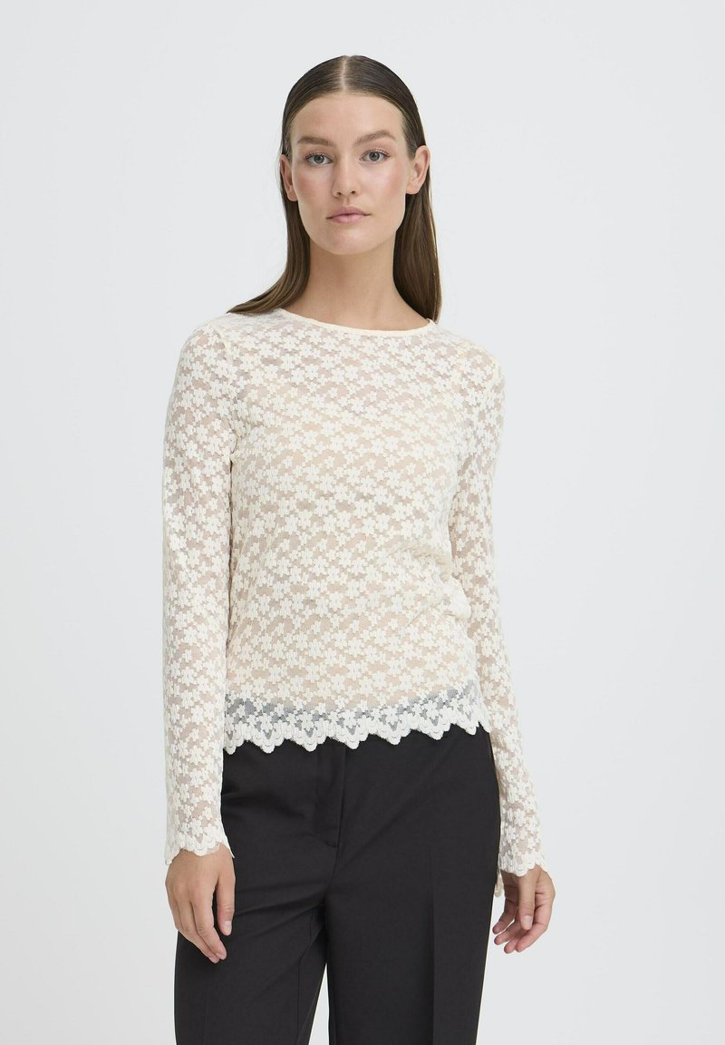 Blonde blonde bluse med lange ærmer i cremefarvet med blomstermønster, bølget kant og en semi-gennemsigtig tekstur, parret med sorte bukser.